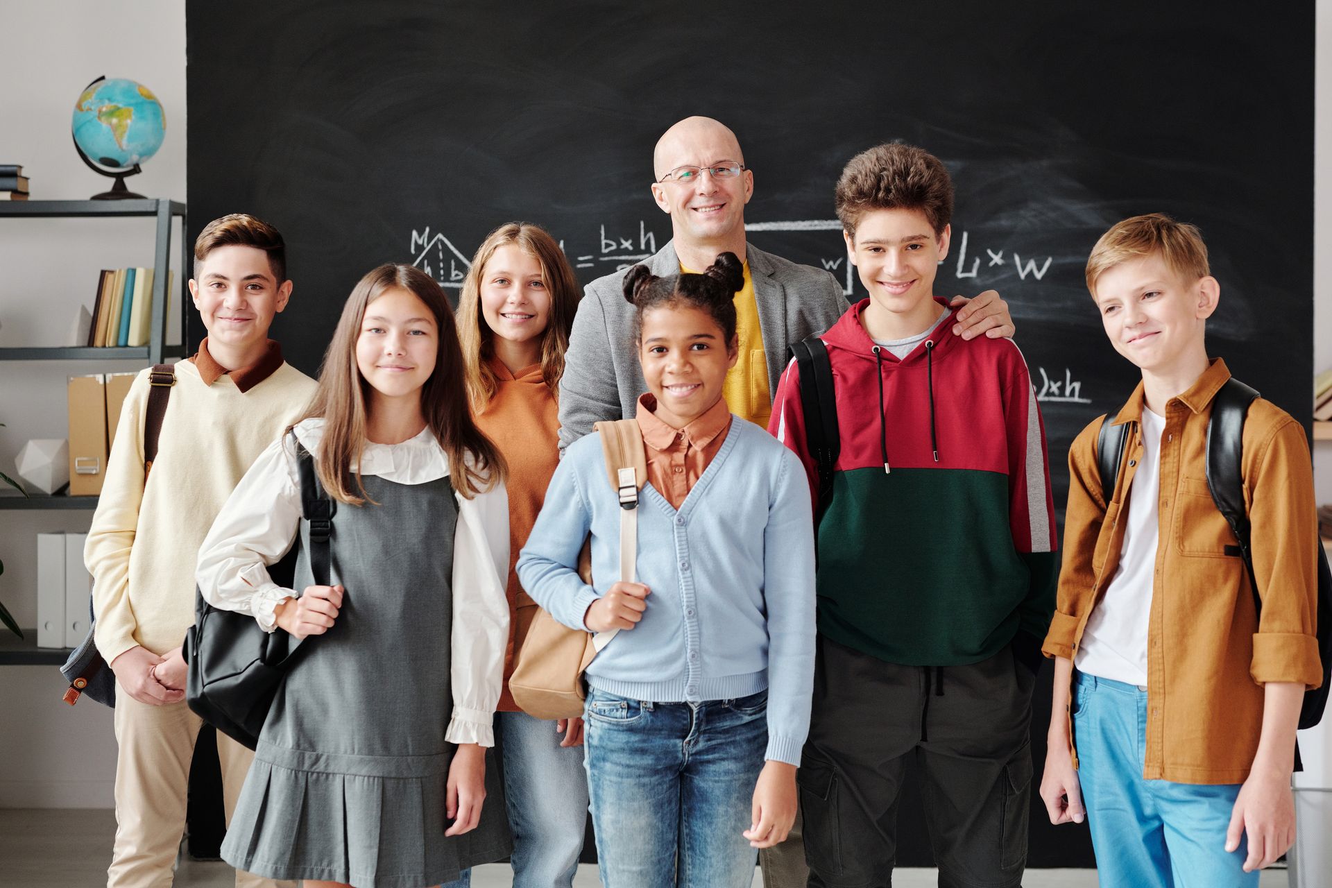 A group of diverse teenagers with backpacks and a teacher smiling in a classroom.